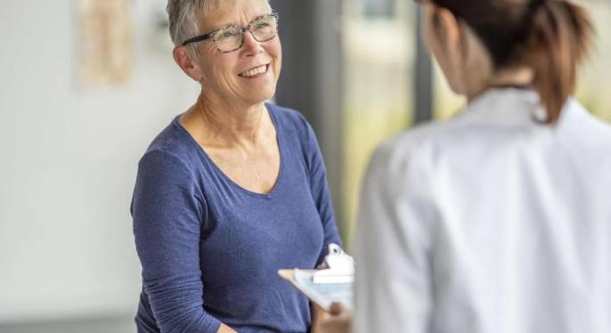 Older woman with short hair and glasses smiling while seated in a doctor office while talking to a doctor in a white coat holding a clip board